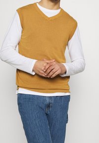 Sennepsgul ærmeløs sweater med V-hals, ribbet kant og glat tekstur, båret over en hvid langærmet skjorte og blå jeans.