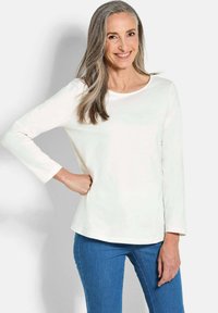 Langärmliges weißes Baumwolloberteil mit rundem Ausschnitt, kombiniert mit engen blauen Jeans. Der Stoff sieht glatt und leicht dehnbar aus.