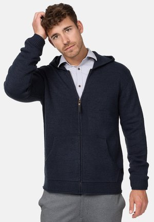 Navy knitted zip-up hoodie with front pockets, ribbed cuffs, and a drawstring hood. Smooth texture and fitted design, paired with a light shirt.