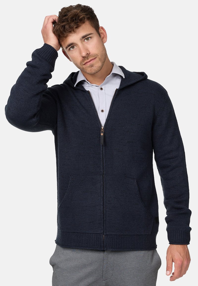 Navy knitted zip-up hoodie with front pockets, ribbed cuffs, and a drawstring hood. Smooth texture and fitted design, paired with a light shirt.
