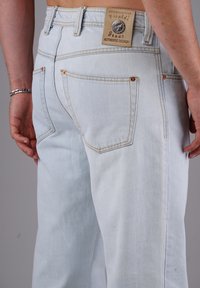 Mann trägt hellblaue Denim-Jeans mit einem Leder-Brandpatch und einem silbernen Knopf am hinteren Bund.