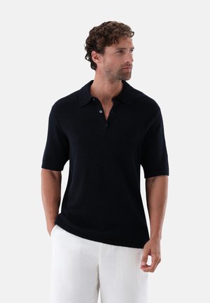 Mann mit lockigem Haar, der ein schwarzes kurzärmeliges Hemd mit Kragen und weiße strukturierte Hose trägt, schaut seitlich mit Händen in den Taschen.