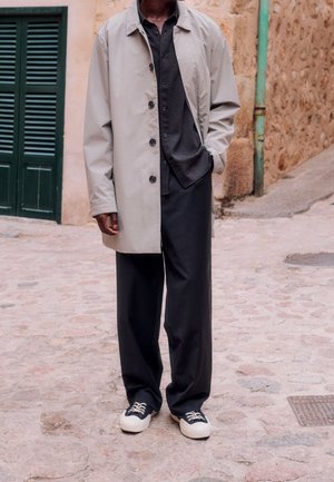 Homme portant un manteau beige clair, une chemise sombre boutonnée, un pantalon noir à jambes larges et des baskets noir et blanc debout dans une rue pavée.