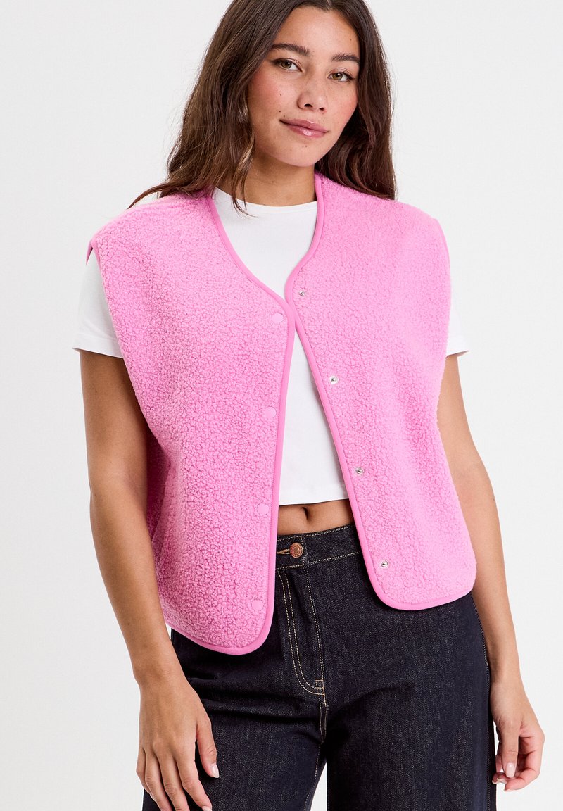 Fleece pink vest with a rounded neckline and snap buttons, featuring a textured surface and contrasting trim. Worn over a white shirt.
