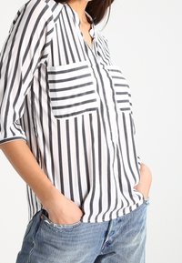 Camicia a righe bianca e nera con placca dei bottoni, due tasche sul petto e maniche a tre quarti. Indossata con jeans in denim.