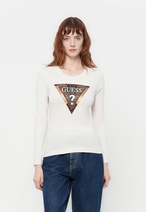 Giovane donna con capelli rossi che indossa una camicia bianca a maniche lunghe con un logo Guess in stampa leopardata metallica e jeans di denim blu.