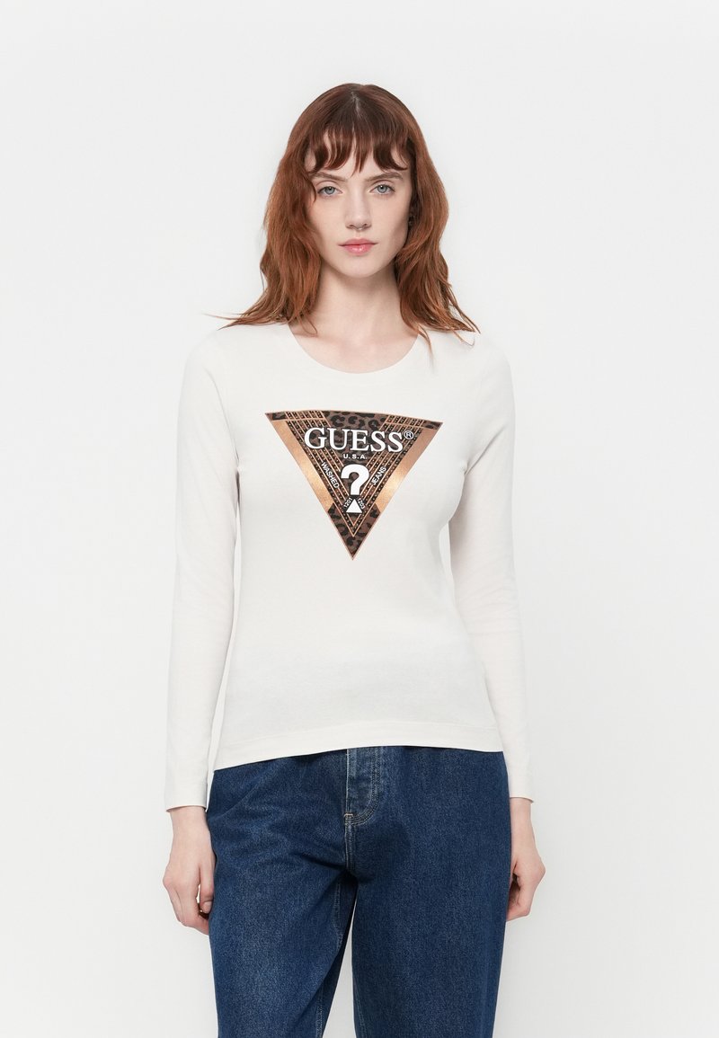 Ung kvinde med rødt hår iført en hvid langærmet skjorte med et metallisk leopardprint Guess-logo og blå denim jeans.