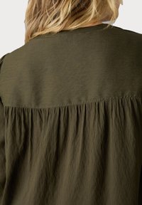 Blouse vert olive avec une texture douce, dotée d'un panneau arrière froncé et d'un empiècement d'épaule lisse. Tissu léger avec une coupe décontractée.