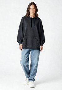 Nő áll, fekete, túlméretezett kapucnis pulóvert visel, elől zsebes, világoskék, bő szabású farmert és fehér tornacipőt visel, egyszínű háttér előtt.