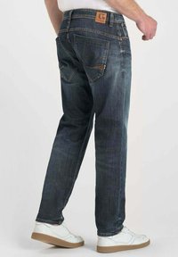 Jeans de mezclilla azul oscuro con corte recto, con bolsillos traseros y un parche de cuero en la cintura, combinados con zapatillas blancas.