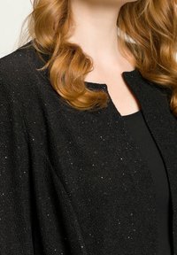 Czarny, fakturowany kardigan z subtelnymi, błyszczącymi akcentami, z okrągłym dekoltem i otwartym przodem, noszony na czarnej bluzce.