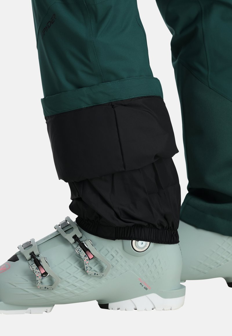 Skihosen aus dunkelgrünem und schwarzem Material mit elastischen Bündchen, kombiniert mit hellgrünen Skischuhen mit verstellbaren Metall-Schnallen und strukturiertem Obermaterial.