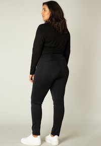 Zwarte fitted leggings met een gladde textuur, zijzakken en een hoge tailleband, gecombineerd met witte sneakers. Achteraanzicht van de outfit.