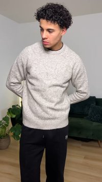 Maglione grigio chiaro con motivo a maglia strutturata, scollo rotondo e maniche lunghe. Indossato con pantaloni jogger neri; sullo sfondo piante e un divano verde.