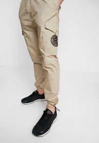 Beige Cargohose mit elastischen Bündchen, ausgestattet mit einer Knie-Tasche und einem schwarzen runden Aufnäher. Getragen mit schwarzen Sneakers, die ein strukturiertes Stoffmuster zeigen.