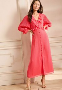 Abito midi corallo plissè con scollatura ruffle, lunghe maniche a palloncino e bottoni frontali. Il tessuto ha una texture liscia e fluida.