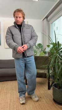 Grijze donzen jas met een hoge kraag en rits, gecombineerd met lichtblauwe jeans. Heeft een gladde textuur en wordt gedragen met sneakers.