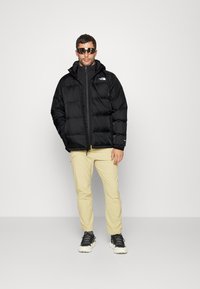 Czarna kurtka puchowa z kapturem, z zapięciem na zamek i logo na piersi. Połączona z jasnobeżowymi spodniami i czarnymi butami sportowymi.