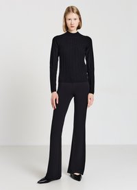 Eine Frau steht vor einem schlichten Hintergrund und trägt einen figurbetonten schwarzen Rippensweater, schwarze weit geschnittene Hosen und schwarze spitze Schuhe.