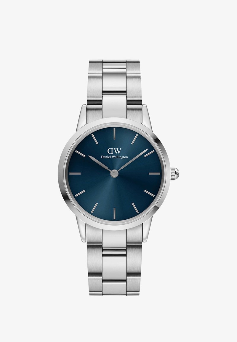 Nerūsējošā tērauda pulkstenis ar apaļu korpusu, zilu saulrieta ciparnīcu, sudraba stundu marķieriem un pulētu metāla siksnu. Mehāniskā pulksteņa kronītis sānos.