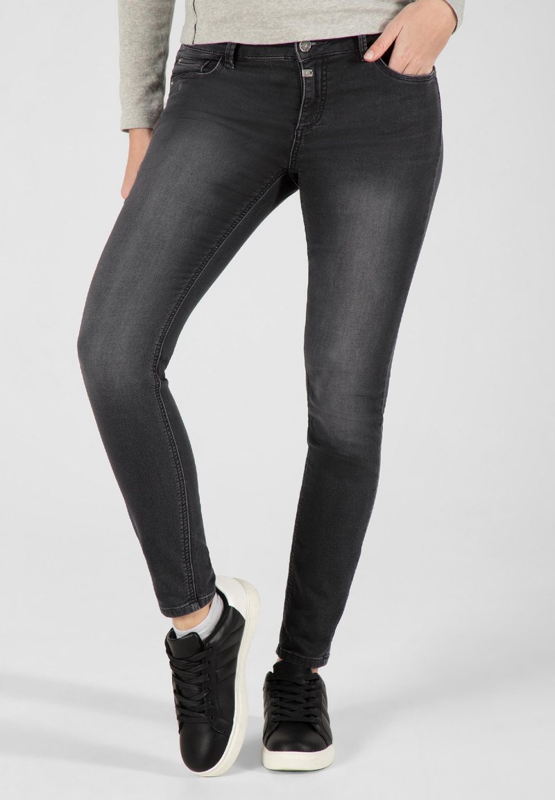 Schwarze Skinny-Jeans mit glatter Textur, ausgestattet mit Fronttaschen, Gürtelschlaufen und Knopfverschluss, kombiniert mit schwarzen Sneakern.
