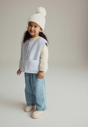 Gilet azzurro chiaro e peloso con chiusura a bottoni e tasche frontali, abbinato a una camicia beige con maniche lunghe, jeans di jeans larghi e stivali color crema.