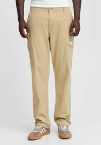 Beige Cargo-Hosen mit zwei Seitentaschen, geradem Bein und Gürtelschlaufen, kombiniert mit braunen Sportschuhen mit weißen Streifen.