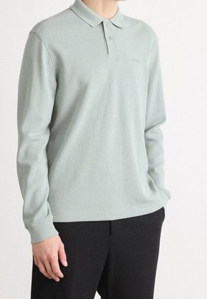 Mężczyzna ubrany w jasnoszarą koszulkę polo z długim rękawem i dwoma guzikami oraz czarne spodnie, stojący z jedną ręką w kieszeni.