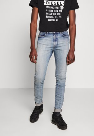 Personne portant un jean slim bleu clair retroussé aux chevilles, des baskets noires et un t-shirt noir à motif Diesel, debout sur un fond uni.