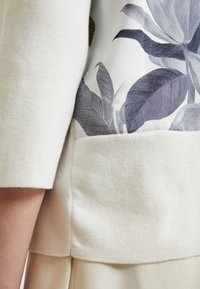 Detail zblízka na rukáv a lem svetru v krémové barvě s potištěnou textilní panelou se širokými tmavě šedými listy.