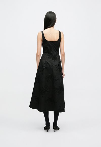 Μαύρο σατέν maxi φόρεμα με εφαρμοστό κορσέ, τετράγωνη λαιμόκοψη και κεντημένα λουλουδάτα σχέδια διακοσμημένα με χάντρες. Φορεμένο με μαύρες καλσόν και τακούνια.