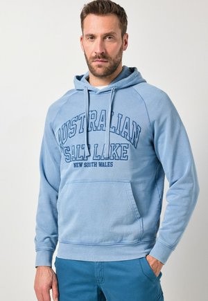 Homme barbu portant un sweat à capuche bleu clair avec le texte « AUSTRALIAN SALT LAKE NEW SOUTH WALES », associé à un pantalon bleu, debout devant un fond uni.