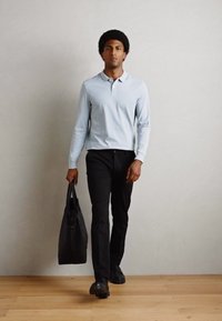 Polo shirt à manches longues bleu clair, pantalon noir et chaussures foncées. Porte un sac fourre-tout noir. Design simple et ajusté avec un minimum de détails.