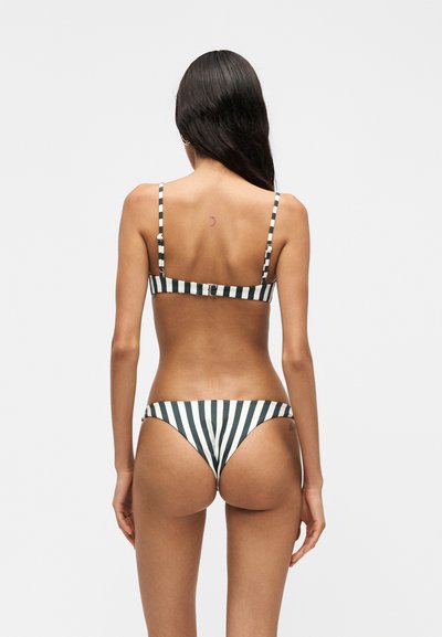 Femme aux longs cheveux foncés vue de dos, portant un bikini rayé verticalement noir et blanc avec des fines bretelles et un bas taille basse.