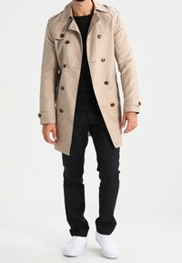 Beżowy trencz z podwójnym rzędem guzików, pagonami na ramionach i kołnierzem z klapami, noszony na czarnej koszuli, w połączeniu z czarnymi spodniami i białymi trampkami.