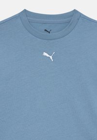 Modrá crewneck tričko s malým bielym logom Puma umiestneným pod golierom a menším čiernym logom Puma vo vnútri goliera.