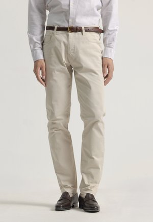 Pantalones de algodón beige con corte slim, que cuentan con un cinturón y bolsillos laterales, combinados con mocasines de piel oscuros. Textura suave, sin patrones.