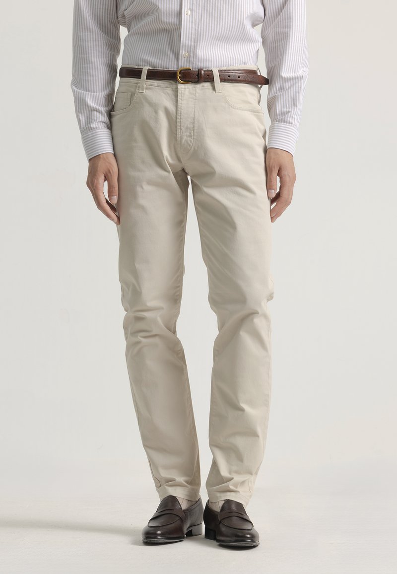 Pantalones de algodón beige con corte slim, que cuentan con un cinturón y bolsillos laterales, combinados con mocasines de piel oscuros. Textura suave, sin patrones.