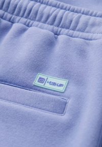 Pantaloni sportivi viola chiaro con una vita testurizzata e una tasca cucita, caratterizzati da un'etichetta rettangolare blu con "LEVEL UP" in bianco.