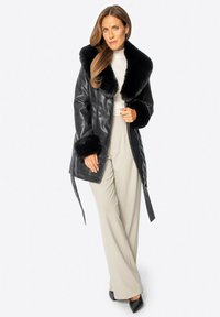 Schwarzer Lederrock mit breitem Pelzkragen und -manschetten. Besitzt einen geraden Schnitt, wird an der Taille gebunden und wird mit beigen weit geschnittenen Hosen kombiniert.