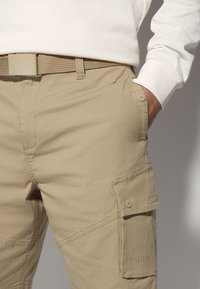 Beige cargo pants made of textured fabric, featuring a side pocket with button closure, and complemented by a matching woven belt.