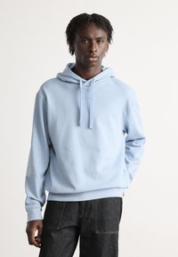 Sudadera azul claro con un bolsillo canguro en el frente, cordón ajustable y puños de canalé, combinada con jeans oscuros. Textura de tela suave.