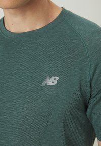 Primer plano de una persona que lleva una camiseta deportiva de New Balance perforada de color verde, con mangas cortas y cuello redondo.