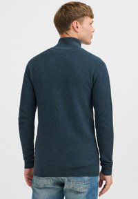 Dark blue knitted sweater with a high collar and long sleeves, featuring a textured pattern, paired with light blue denim jeans.