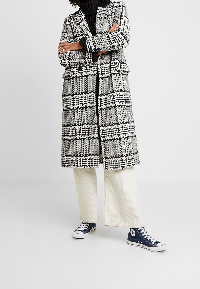 Manteau long à carreaux en noir, blanc et gris, avec un design croisé à double boutonnage. Porté sur un col roulé noir et associé à un pantalon large crème et des baskets.