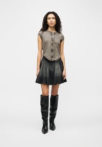 Γυναίκα με σγουρά μαλλιά, φορεμένη με γκρι αμάνικο πουκάμισο με κουμπιά, μαύρη φούστα με καμπάνα και μαύρες δερμάτινες μπότες μέχρι το γόνατο, στέκεται μπροστά σε απλό φόντο.