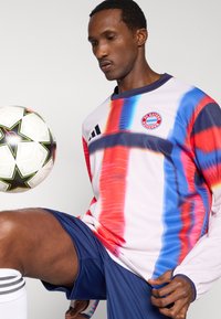 Ilgas rankovėmis, margas futbolo marškinėlis su mėlynais, raudonais ir baltais raštais, kuriame yra apskritas „Bayern Munchen“ logotipas ir juodos petnešėlės.