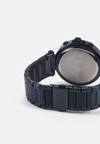 Reloj de pulsera negro de metal con una correa gruesa y acabado pulido. La caja redonda presenta botones de cronógrafo y una parte trasera suave con el logo de la marca.