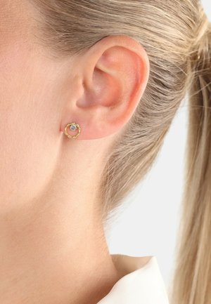 Orecchini a cerchio in tono oro con un design intrecciato, dotati di un centro in pietra trasparente, indossati all'orecchio di un modello.