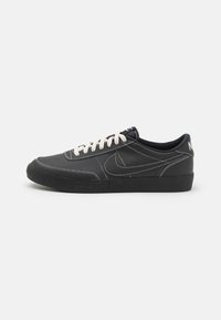 Fekete bőr sportcipők, texturált fekete gumitalppal, fehér varrással és krémszínű fűzővel. Nike logó látható az oldalon.
